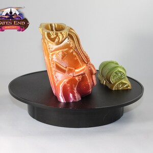 May include: A 3D printed miniature figure of a person wearing a cloak and holding a staff. The figure is painted in shades of orange, red, and yellow. The figure is standing on a black circular base. A smaller 3D printed object, a green and gold rolled-up bag, is on the base to the right of the figure. The text "Fates End MMF547" is visible in the background.