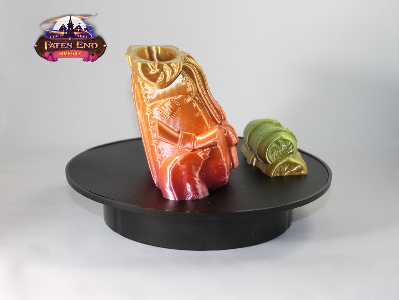 May include: A 3D printed miniature figure of a person wearing a cloak and holding a staff. The figure is painted in shades of orange, red, and yellow. The figure is standing on a black circular base. A smaller 3D printed object, a green and gold rolled-up bag, is on the base to the right of the figure. The text "Fates End MMF547" is visible in the background.