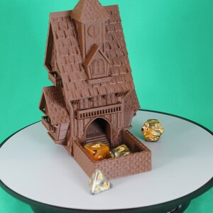 May include: A brown 3D printed dice tower shaped like a house with a pointed roof and a small window. The tower has a drawer that opens to reveal a dice tray. There are four dice inside the tray, three are gold and one is green. The tower is sitting on a black rotating base with a white surface. The text "TINY TOWERS FATES END TT241" is visible on a label in the upper right corner of the image.
