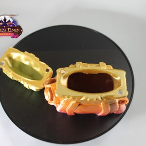 May include: A gold and orange treasure chest with a hinged lid open, revealing an empty interior. The chest has a textured surface and is decorated with raised details.