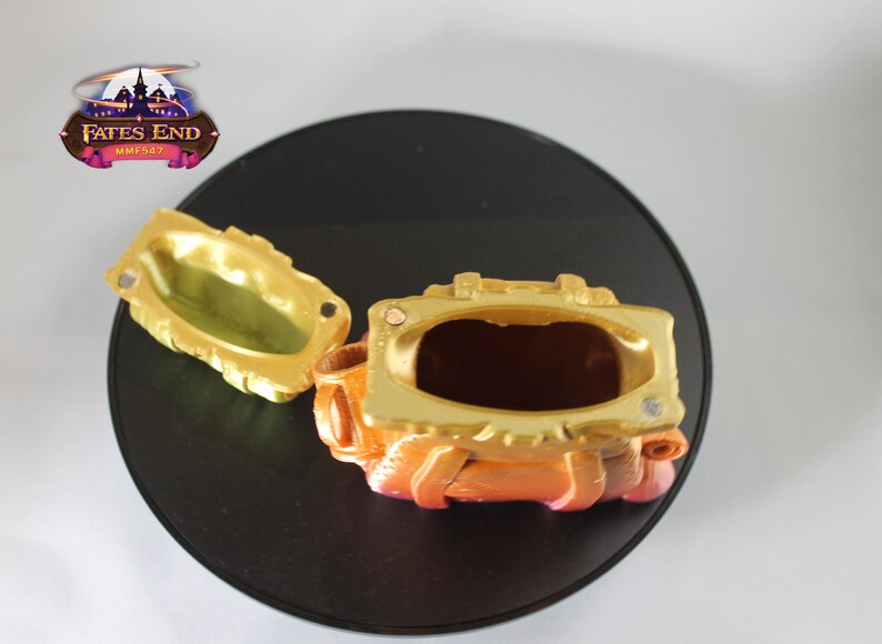 May include: A gold and orange treasure chest with a hinged lid open, revealing an empty interior. The chest has a textured surface and is decorated with raised details.