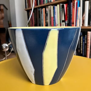 Peut inclure: Un bol en céramique avec un extérieur bleu marine, orné de rayures verticales blanches et jaunes. L'intérieur est jaune pâle. Le bol est posé sur une surface jaune, avec une bibliothèque en arrière-plan.