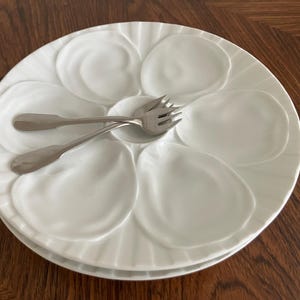 Puede incluir: Un plato para ostras de cerámica blanca con seis hendiduras moldeadas en forma de concha dispuestas en un patrón circular alrededor de un hueco central. Dos tenedores para marisco de acero inoxidable de tres púas descansan en el centro.