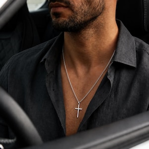 May include: A silver-toned metal cross pendant suspended from a matching thin cable chain necklace. The cross features a simple, clean design with straight edges and a polished finish, resting against a dark charcoal gray button-down shirt.