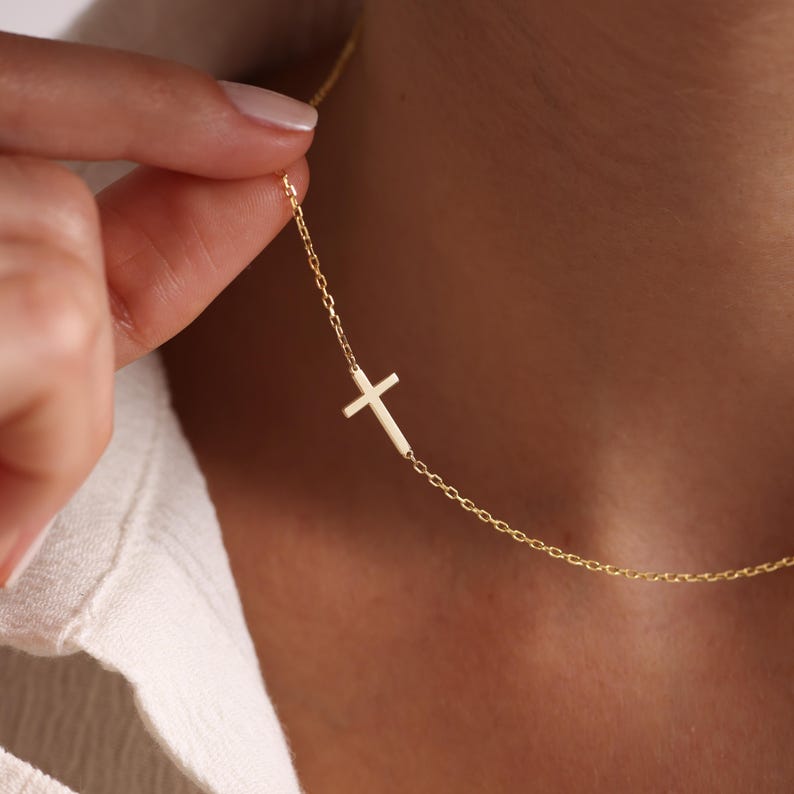 May include: A delicate gold-colored necklace featuring a sideways cross pendant. The cross is attached to a fine chain, creating a minimalist and elegant design. The necklace is shown against a skin tone background.