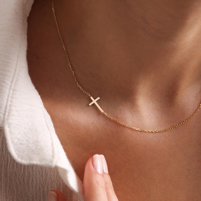 May include: A delicate gold necklace featuring a sideways cross pendant. The cross is attached to a fine chain, creating a minimalist and elegant design. The necklace is shown against a white shirt.