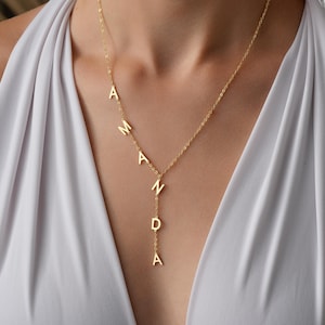 May include: A gold-toned necklace featuring a delicate chain with the name AMANDA spelled out in capital letters. The letters are arranged in a vertical drop design, with the first three letters positioned along the chain and the remaining letters hanging down in a straight line from the center point.
