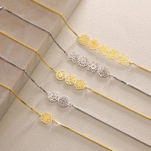 May include: Five delicate gold chains with different floral designs. Each chain features a series of interconnected flower cutouts, creating a delicate and intricate pattern.