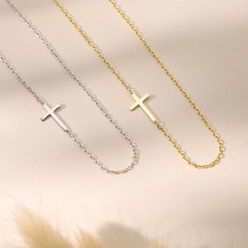 May include: Close-up of three delicate necklaces. One is silver, and the other two are gold. Each necklace features a small cross pendant offset on a delicate chain. The necklaces are displayed on a neutral background.