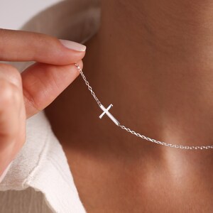 May include: A delicate silver necklace featuring a small cross pendant. The cross is positioned to the side on a fine chain. The necklace is shown against a neutral background, highlighting its simple elegance.