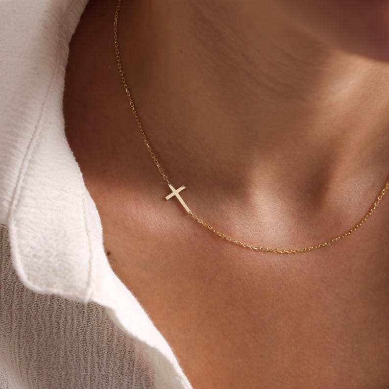 May include: A delicate gold-colored necklace featuring a small cross pendant. The cross is positioned to the side of the chain, creating an asymmetrical design. The necklace is shown against a white shirt.