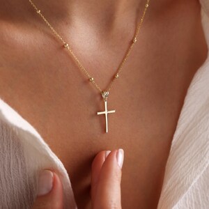 Op de afbeelding: Een gouden ketting met een klein gouden kruisje als hanger. De ketting is gemaakt van kleine, ronde kralen.