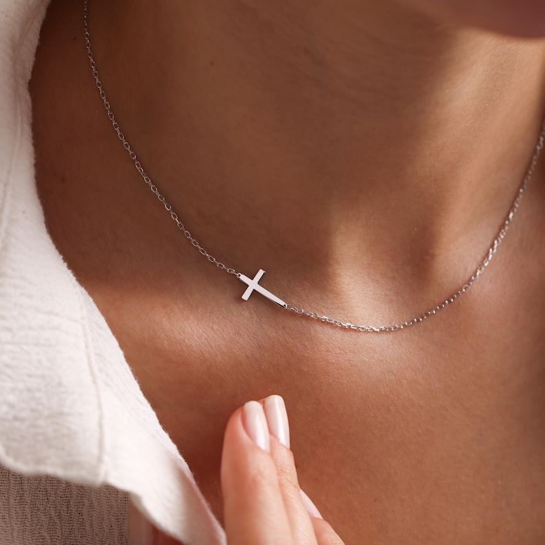 May include: A delicate silver necklace featuring a sideways cross pendant. The cross is positioned to the left, attached to a fine chain. The necklace is worn on a person's neck, against a white shirt.