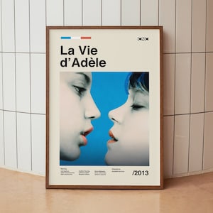 Puede incluir: Un póster de película enmarcado para la película "La Vie d'Adèle" con un fondo azul y un primer plano de dos caras de mujeres. El póster incluye el título de la película, el año 2013 y los nombres de los actores y el director.