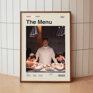 以下が含まれることがあります： レストランのキッチンにいる人々のグループを描いた映画「ザ・メニュー」のポスター。ポスターは茶色の木製の額縁に入っていて、白地に黒文字で書かれている。ポスターのテキストには、映画のタイトル、俳優の名前、監督の名前が含まれている。ポスターには2022年も記載されている。