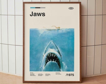 Könnte beinhalten: Ein gerahmtes Poster mit minimalistischem Design, das den Titel "Jaws" in fettem weißen Text zeigt. Das Poster zeigt einen großen Hai mit offenem Maul, der teilweise im blauen Wasser versunken ist. Das Jahr "1975" ist in der rechten unteren Ecke gedruckt.