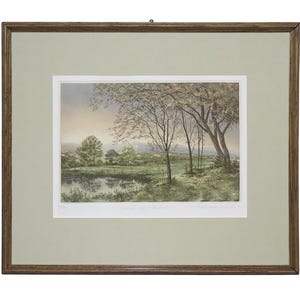 May include: A framed landscape print featuring a pond in the foreground, green fields, and trees under a soft sky. The artwork is enclosed in a light green mount and a dark brown wooden frame, with handwritten text visible at the bottom of the print.