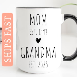 May include: White ceramic coffee mug with a black rim and handle. The mug has black text that reads "Mom Est. 1998" and "Grandma Est. 2025" with a small black heart between the two lines of text.