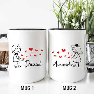 May include: Two white coffee mugs with black handles and black rims. Each mug has a simple black line drawing of a person holding a red heart. The mug on the left says "Daniel" and the mug on the right says "Amanda".
