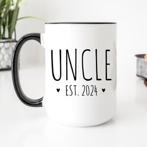 May include: White ceramic coffee mug with a black rim and handle. The mug has black text that reads "UNCLE EST. 2024" with a small heart on either side of the year.