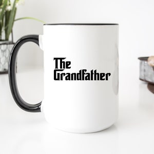 Op de afbeelding: Witte keramische mok met een zwarte rand en handvat. De mok heeft de tekst "The Grandfather" in een vetgedrukte, zwarte lettertype. De mok staat op een wit oppervlak.