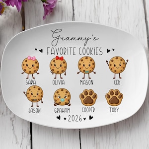 Peut inclure: Assiette ovale en c&eacute;ramique blanche avec le texte "Grammy's Favorite Cookies" et l'ann&eacute;e 2026. L'assiette pr&eacute;sente des personnages de biscuits de dessins anim&eacute;s avec des noms comme Sara, Olivia, Mason et Ced. Les biscuits ont des bras, des jambes et des expressions faciales.