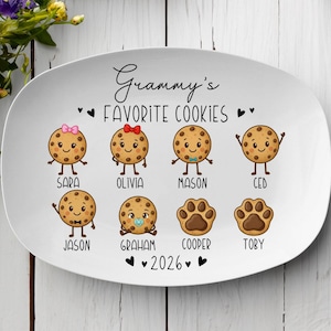 Peut inclure: Assiette ovale en c&eacute;ramique blanche avec le texte "Grammy's FAVORITE COOKIES" et l'ann&eacute;e "2026". L'assiette pr&eacute;sente des biscuits de dessin anim&eacute; avec des noms comme Sara, Olivia, Mason, Ced, Jason, Graham, Cooper et Toby.
