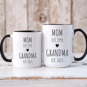 May include: Two white ceramic mugs with black handles and black rims. The mugs have black text that reads "MOM EST. 1998 GRANDMA EST. 2025" on one mug and "MOM EST. 1998 GRANDMA EST. 2025" on the other mug.