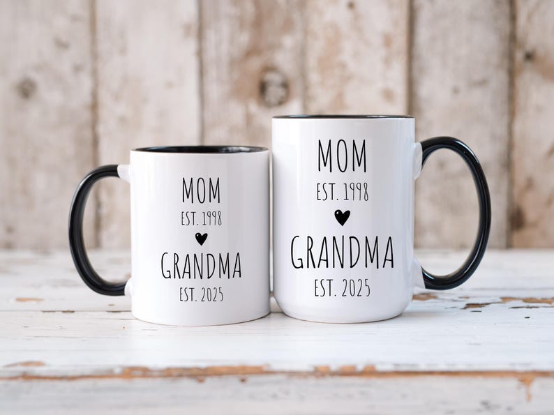 May include: Two white ceramic mugs with black handles and black rims. The mugs have black text that reads "MOM EST. 1998 GRANDMA EST. 2025" on one mug and "MOM EST. 1998 GRANDMA EST. 2025" on the other mug.