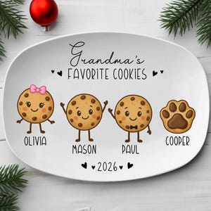 Peut inclure: Assiette ovale en c&eacute;ramique blanche avec le texte "Grandma's Favorite Cookies". L'assiette pr&eacute;sente des biscuits de dessin anim&eacute; avec les noms Olivia, Mason, Paul et Cooper. Un n&oelig;ud rose orne le biscuit d'Olivia. L'ann&eacute;e 2026 est &eacute;galement affich&eacute;e.