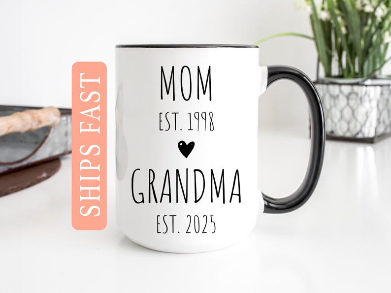 May include: White ceramic coffee mug with a black rim and handle. The mug has black text that reads "Mom Est. 1998" and "Grandma Est. 2025" with a small black heart between the two lines of text.