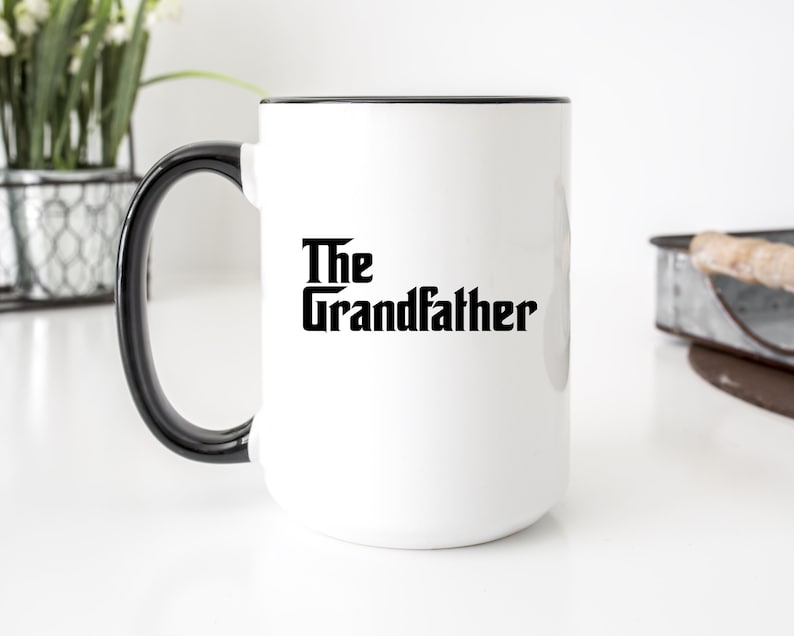 Op de afbeelding: Witte keramische mok met een zwarte rand en handvat. De mok heeft de tekst "The Grandfather" in een vetgedrukte, zwarte lettertype. De mok staat op een wit oppervlak.