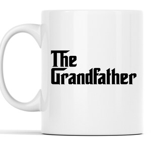Op de afbeelding: Witte keramische mok met een gebogen oor. De mok heeft de woorden "The Grandfather" in vetgedrukte, zwarte, gestileerde letters. Het ontwerp is gecentreerd op het oppervlak van de mok. De mok heeft een standaardformaat voor warme dranken.