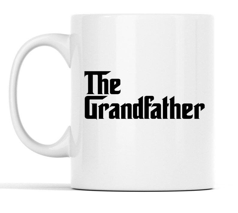 Op de afbeelding: Witte keramische mok met een gebogen oor. De mok heeft de woorden "The Grandfather" in vetgedrukte, zwarte, gestileerde letters. Het ontwerp is gecentreerd op het oppervlak van de mok. De mok heeft een standaardformaat voor warme dranken.