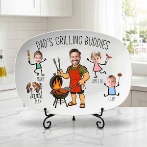 Peut inclure: Un plat ovale blanc avec le texte "DAD'S GRILLING BUDDIES" et des illustrations de dessins anim&eacute;s d'un homme grillant, de trois enfants et d'un chien. L'homme porte un tablier et tient des ustensiles de cuisine. Le plat est sur un support en m&eacute;tal noir.