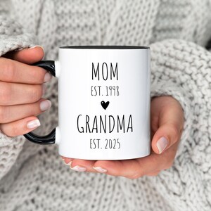 May include: White ceramic mug with a black handle and black rim. The mug has black text that reads "MOM EST. 1998" with a small black heart and "GRANDMA EST. 2025".