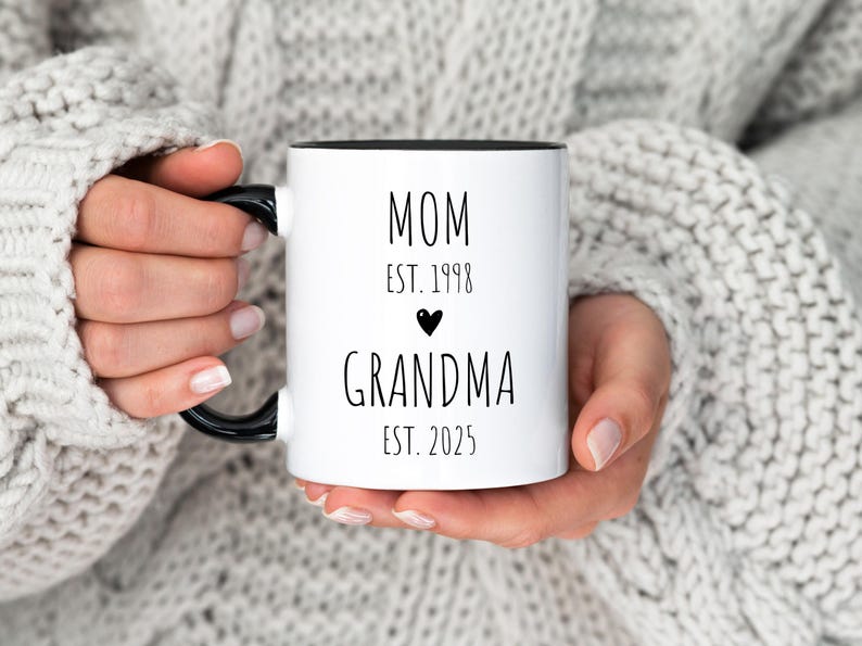 May include: White ceramic mug with a black handle and black rim. The mug has black text that reads "MOM EST. 1998" with a small black heart and "GRANDMA EST. 2025".