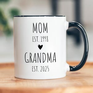 May include: White ceramic coffee mug with a black rim and handle. The mug has black text that reads "MOM EST. 1998" and "GRANDMA EST. 2025" with a small black heart between the two lines of text.