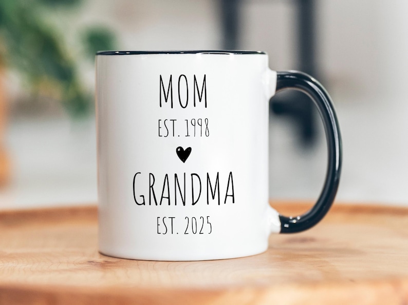 May include: White ceramic coffee mug with a black rim and handle. The mug has black text that reads "MOM EST. 1998" and "GRANDMA EST. 2025" with a small black heart between the two lines of text.