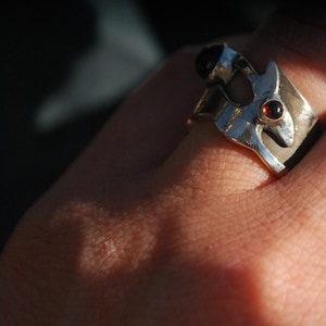 Könnte beinhalten: Ein silberner Ring mit einem stilisierten Tierdesign und einem roten Edelstein. Der Ring wird an einem Finger getragen.