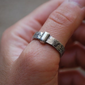 May include: A silver ring with a textured band and a rectangular silver accent. The ring is worn on a person's finger.