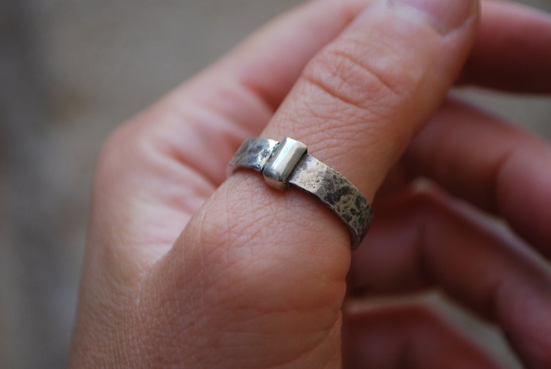May include: A silver ring with a textured band and a rectangular silver accent. The ring is worn on a person's finger.