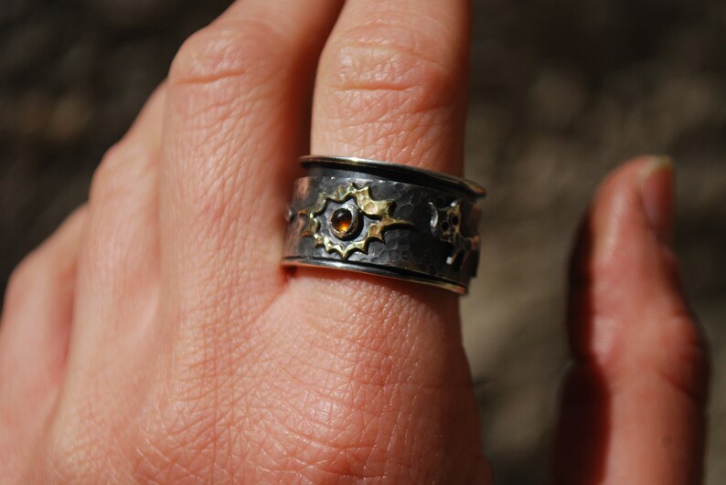 May include: A silver and gold ring with a hammered finish and a small amber gemstone. The ring is decorated with a stylised design of stars and other geometric shapes.