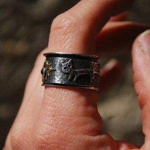 May include: A silver ring with a hammered finish and a gold cat design. The ring is worn on a person's finger.