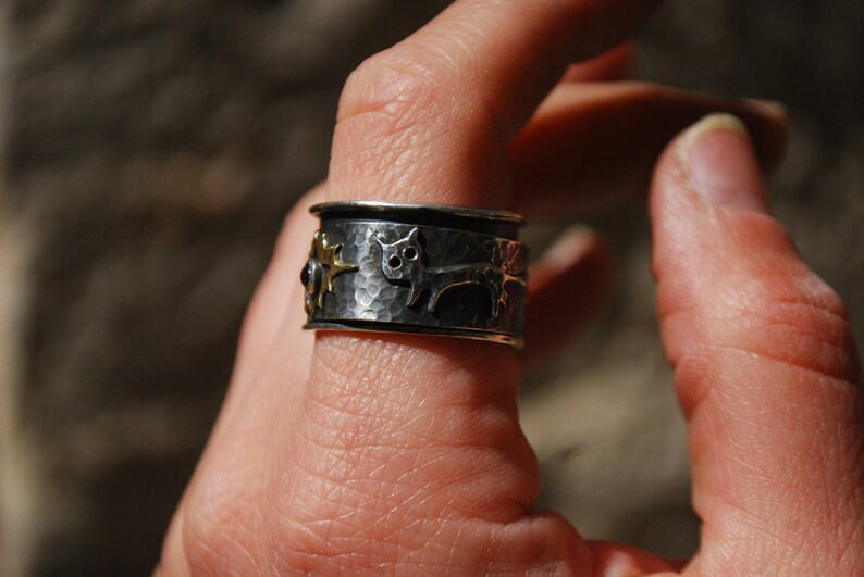 May include: A silver ring with a hammered finish and a gold cat design. The ring is worn on a person's finger.