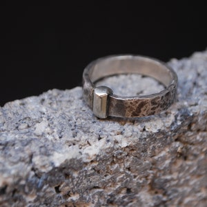 May include: A silver ring with a hammered texture and a gold accent. The ring is sitting on a rough, gray surface.