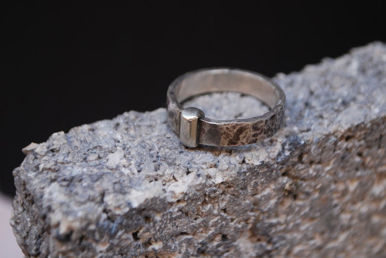 May include: A silver ring with a hammered texture and a gold accent. The ring is sitting on a rough, gray surface.