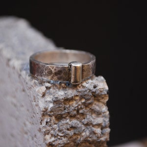 May include: A silver ring with a textured finish and a rectangular band. The ring is sitting on a rough, gray surface.