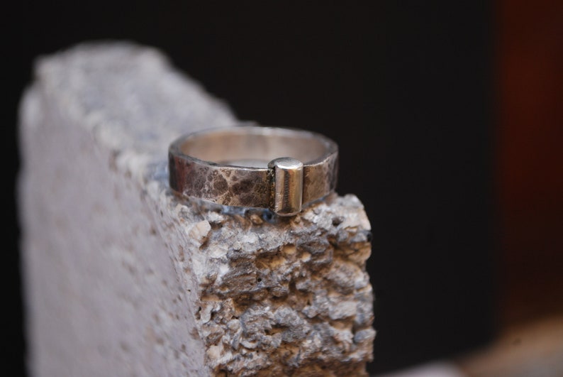 May include: A silver ring with a textured finish and a rectangular band. The ring is sitting on a rough, gray surface.