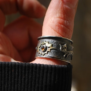 May include: A silver and gold ring with a hammered texture and a small orange gemstone. The ring features a stylised animal design.
